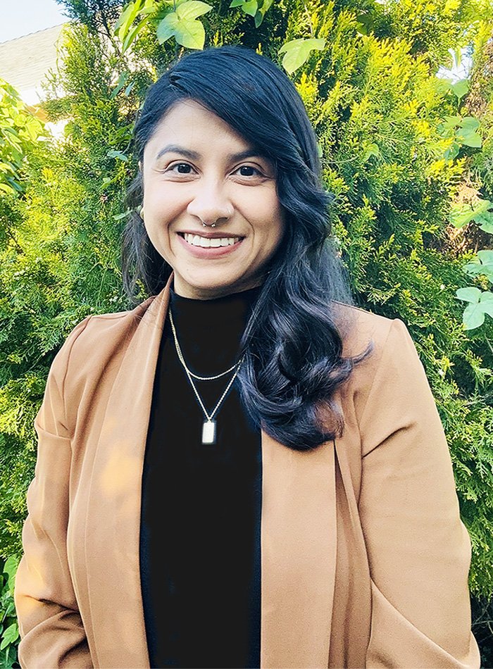 Therapist smiling at camera wearing tan blazer against green foliage backdrop